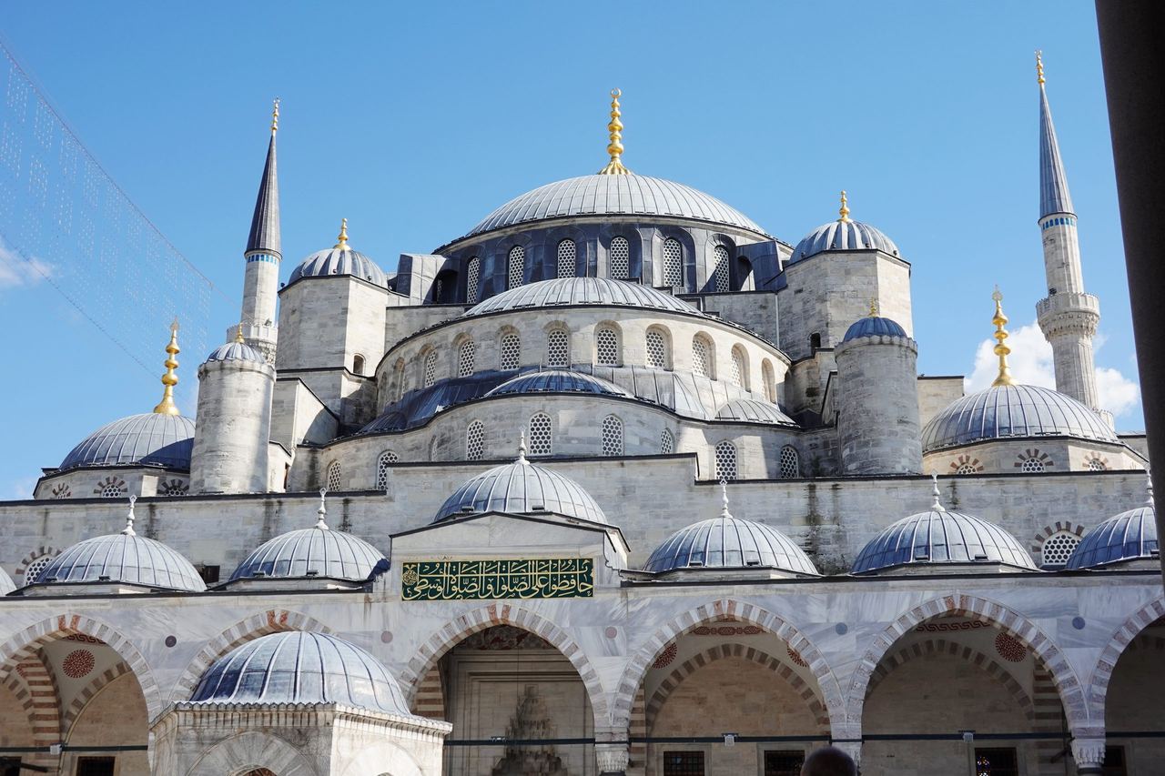 Istanbul’s Blue Mosque fills the frame with cascading gray domes, slender minarets, and a courtyard arcade beneath a clear blue sky, conveying calm grandeur.
