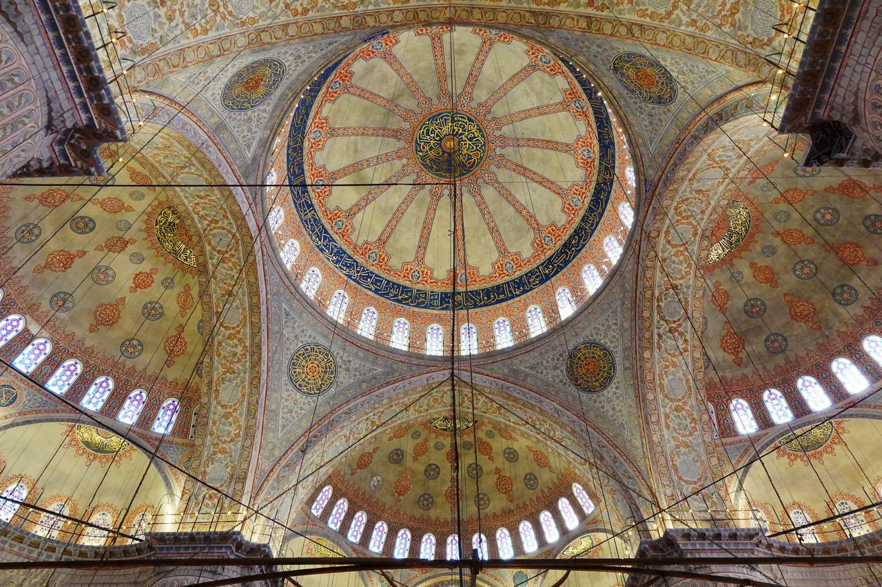 Blue Mosque's interior viewed upward, with a vast central dome, smaller domes, arched windows, and warm gold-and-blue ornamentation.