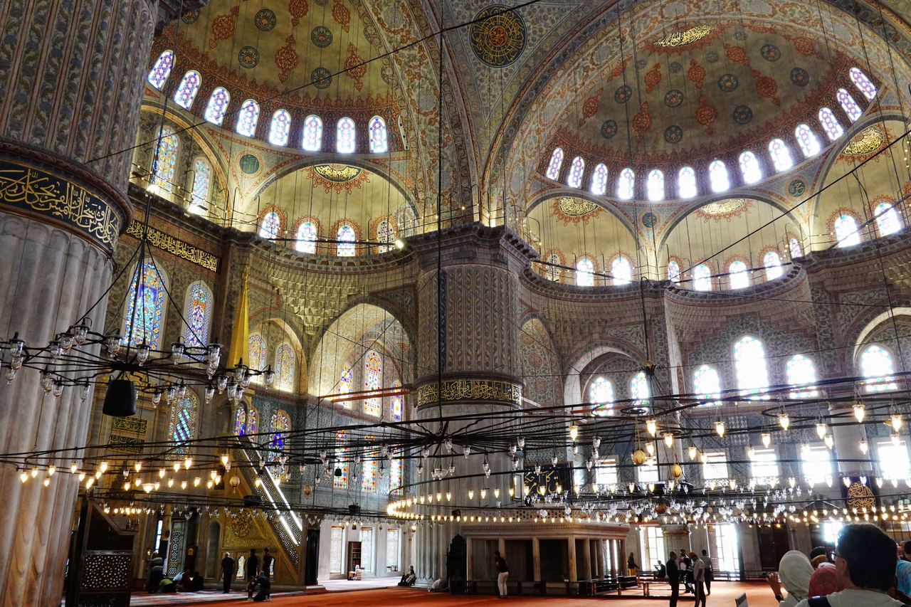 Ornate Blue Mosque's interior with soaring painted domes, arched stained-glass windows, calligraphy bands, and glowing lamps; visitors below.