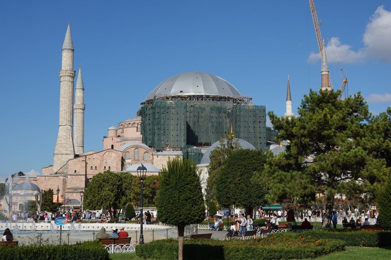 Hagia Sophia in Istanbul rises behind trees, with its central dome under restoration scaffolding, slender minarets, and visitors in the sunny plaza.