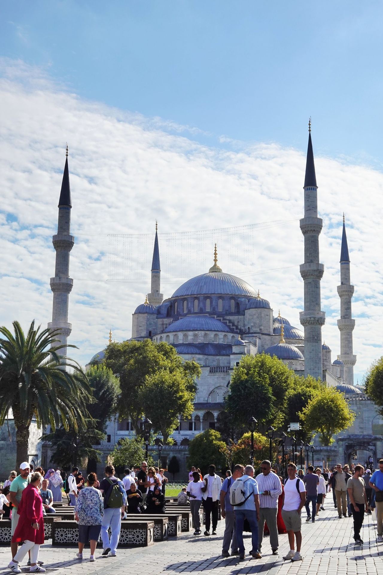 The Blue Mosque’s domes and slender minarets rise above a busy Istanbul plaza, where visitors gather beneath a bright, cloud-streaked sky.