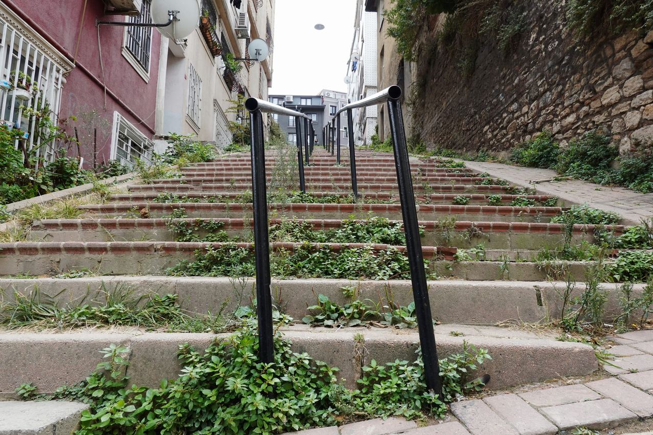 Overgrown stone steps climb between colorful apartment buildings and a rough stone wall, with black handrails guiding the quiet, worn stairway.