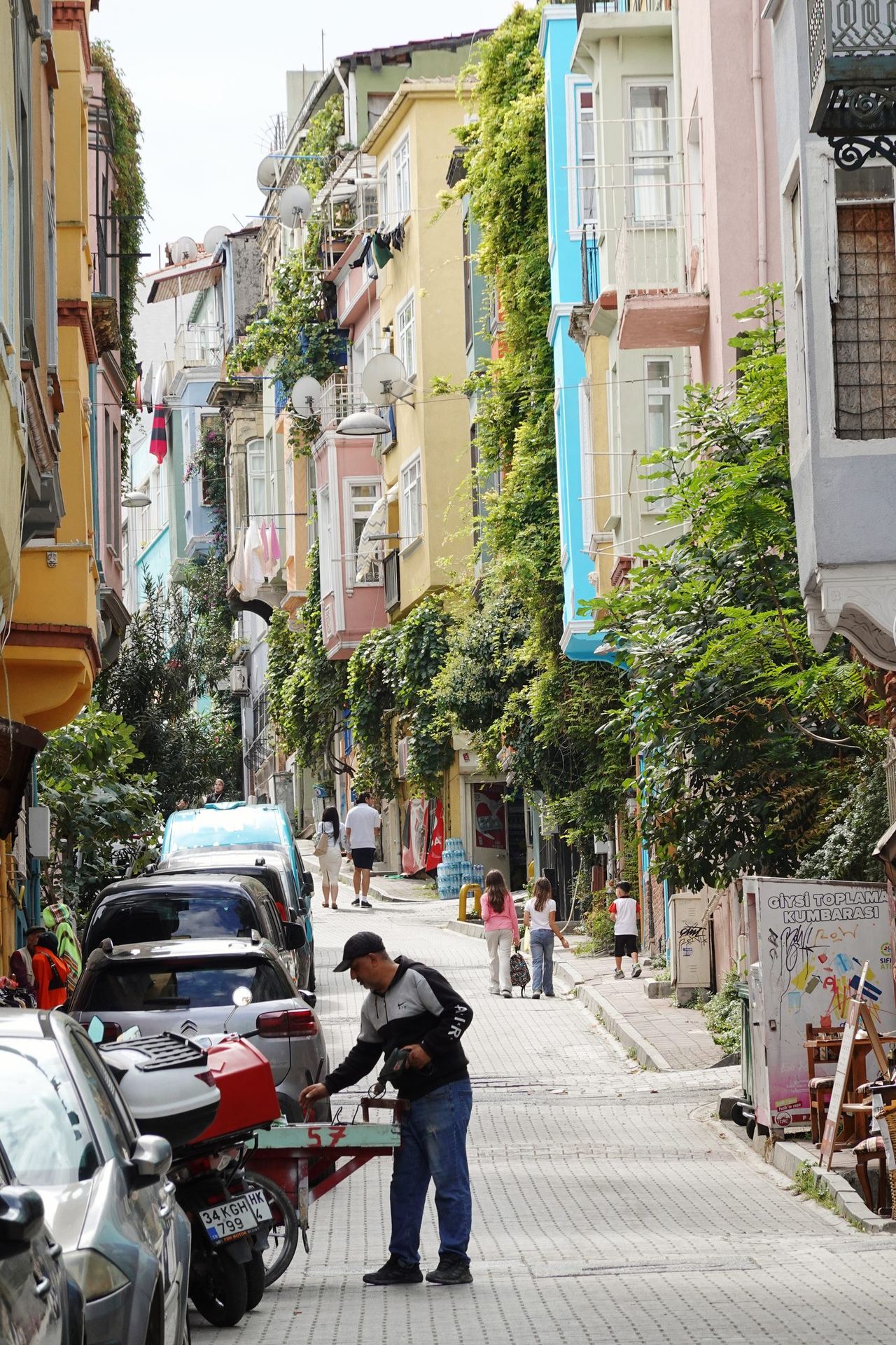 Kiremit street lined with colorful homes, balconies, and greenery; pedestrians pass parked cars as a man works beside a scooter.
