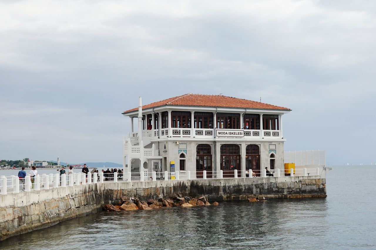 White, red-tiled Moda pier, with visitors along the railings and calm gray water beneath an overcast sky.