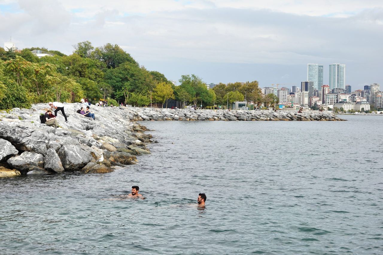 Swimmers cool off in calm waters of Moda beside a rocky, tree-lined shore, with city high-rises in the distance under a bright, cloudy sky.
