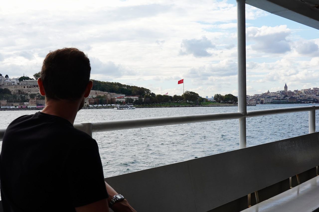 A man in a black shirt looks across the water from a ferry, toward Istanbul’s skyline and a Turkish flag under a cloudy sky; calm, reflective mood.