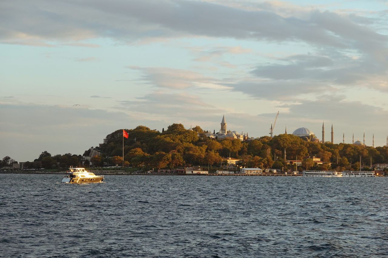 A small boat crosses the rippling Bosphorus before Istanbul’s tree-covered skyline, minarets, domes, and Turkish flag under a calm, pastel evening sky.