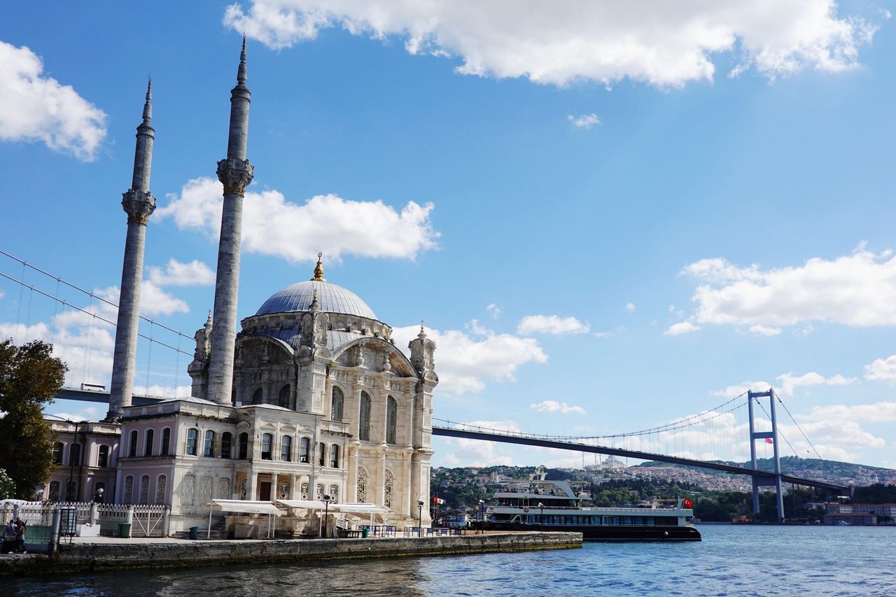 Ortaköy Mosque with its dome and two minarets stands beside the Bosphorus in Istanbul, with a ferry and suspension bridge under a bright blue sky.