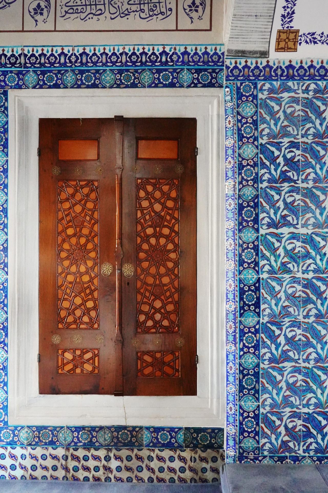 Ornate wooden double window with intricate latticework sit in a white stone frame, surrounded by vivid blue Islamic tiles and Arabic calligraphy.