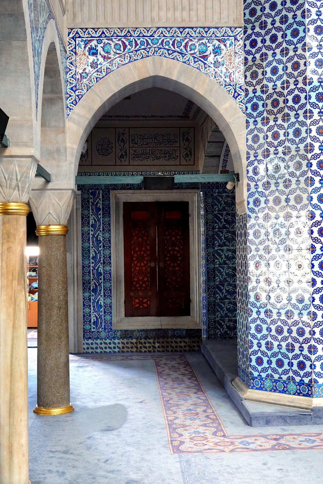 Ornate mosque window with a pointed stone arch, blue-and-white floral tilework, gold-trimmed columns, and a dark wooden door; serene and historic.