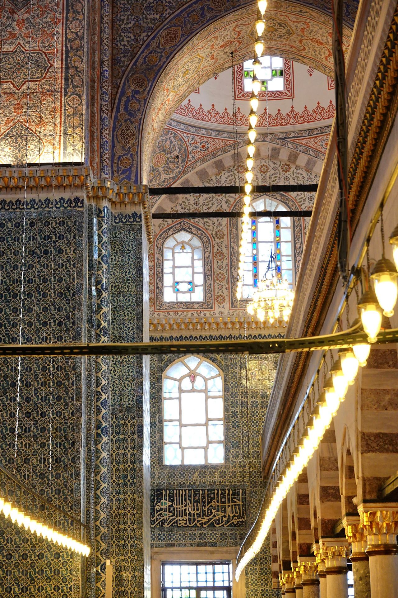 Ornate Rustem Pasha Mosque interior glows with golden chandeliers beneath painted arches and colorful stained-glass windows, blue tile, and Arabic calligraphy.
