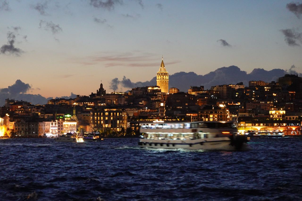 A ferry crosses dark water before Istanbul’s illuminated Galata Tower and hillside skyline at dusk, with warm city lights creating a serene evening mood.