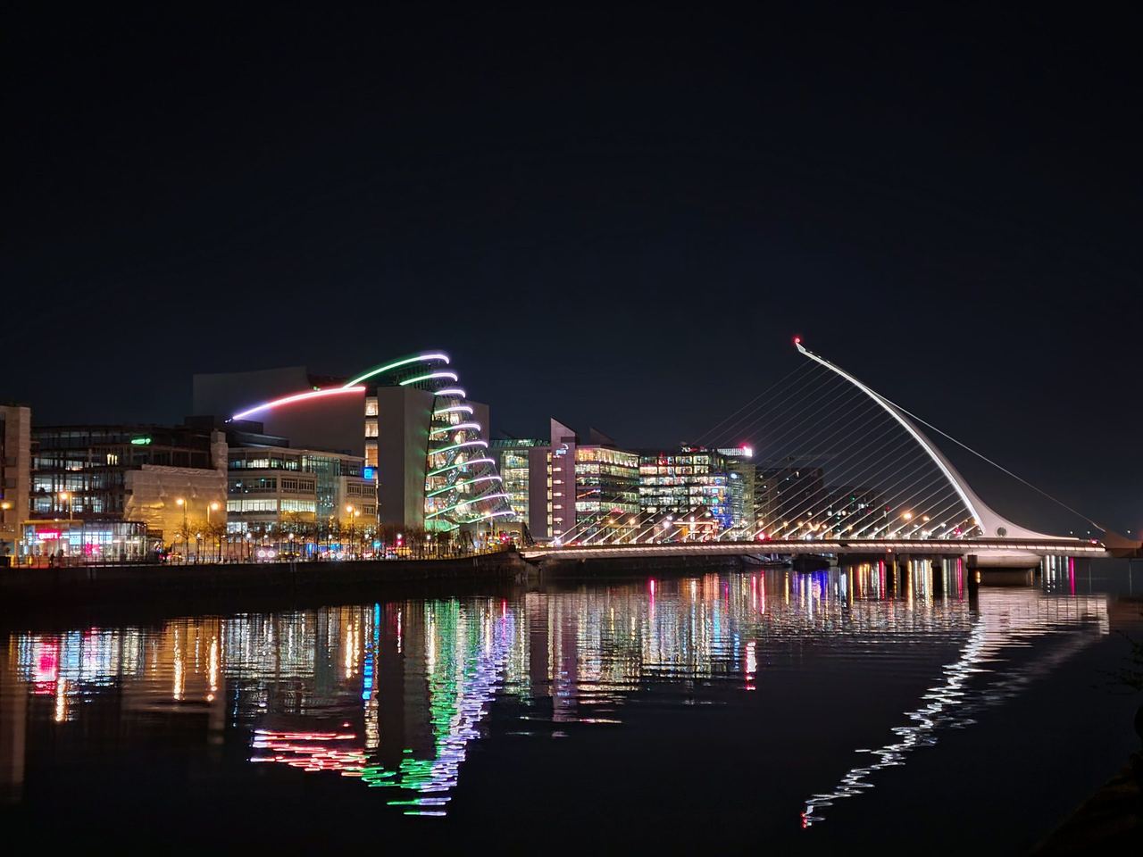 Night view of a city from a river shore with a modern suspension bridge and line of modern office buildings