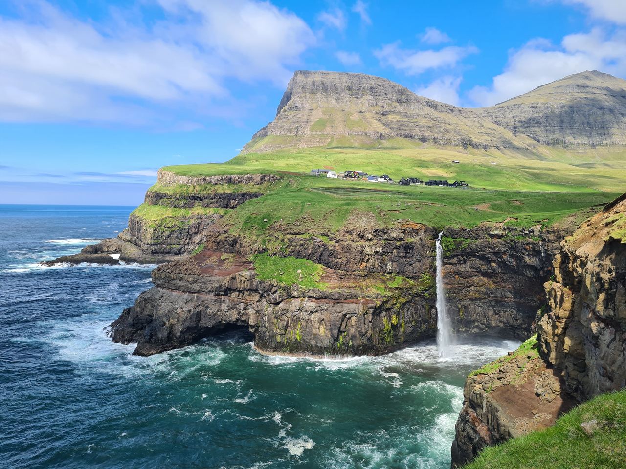 A waterfall dropping straight from a green clifftop into the sea, with a handful of houses on the plateau above